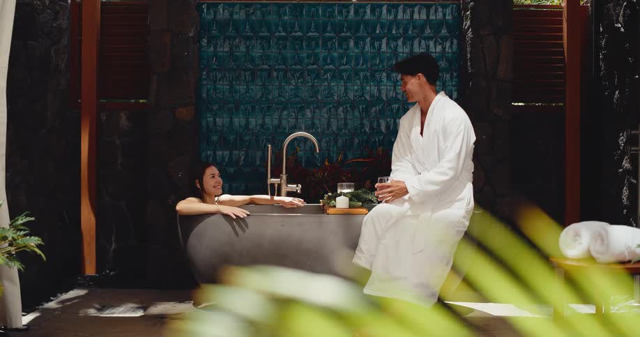 A happy couple enjoys a relaxing spa day together as the woman soaks in an outdoor bathtub and her partner in a bathrobe sits beside her