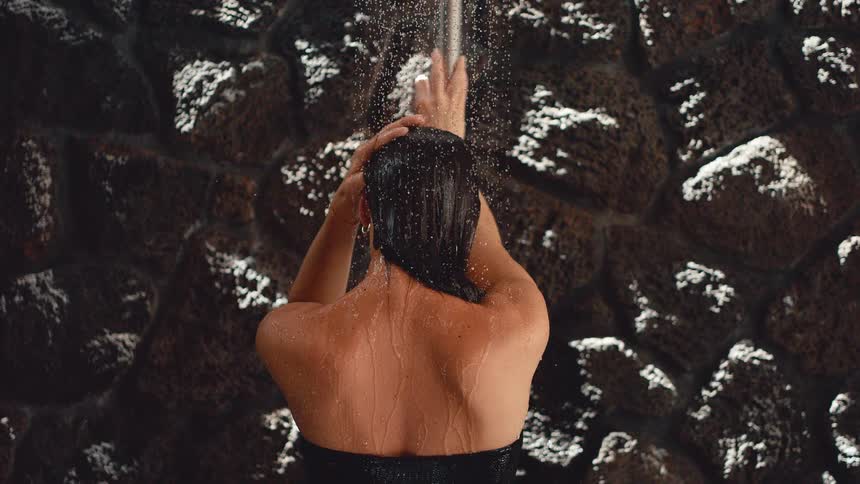 Woman with her back to the camera takes a relaxing shower against a stone wall in slow motion