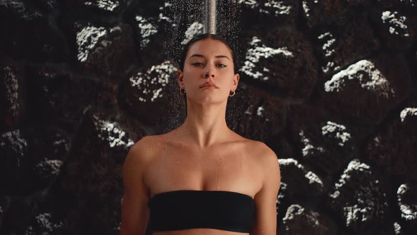 Cinemagraph loop of a serene woman in a black bikini top meditating under a shower in slow motion
