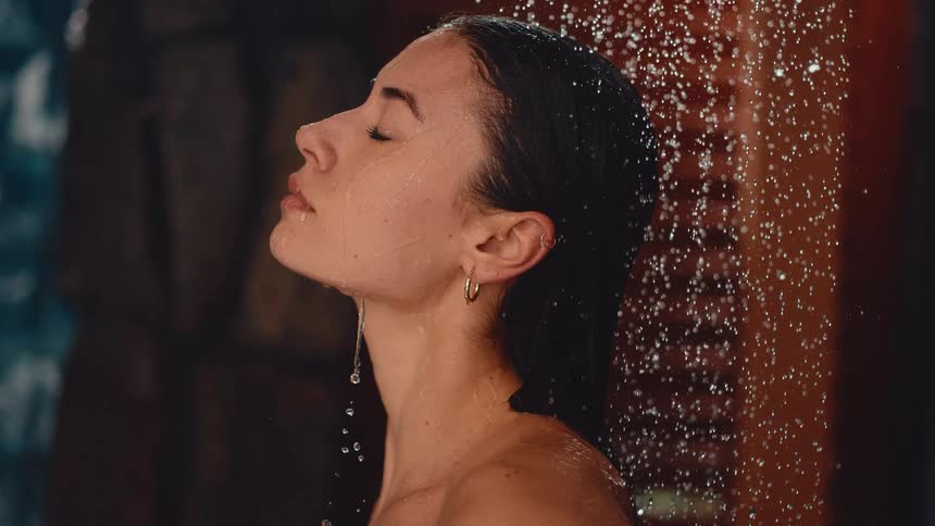 A serene woman with her eyes closed enjoys a relaxing shower as water falls on her face in slow motion