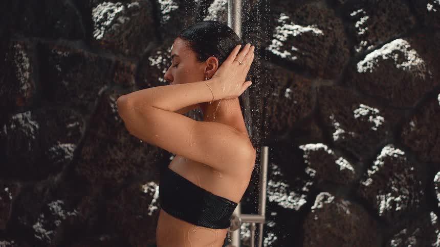 Slow motion cinemagraph of a beautiful woman with her eyes closed enjoying a refreshing shower against a dark stone wall