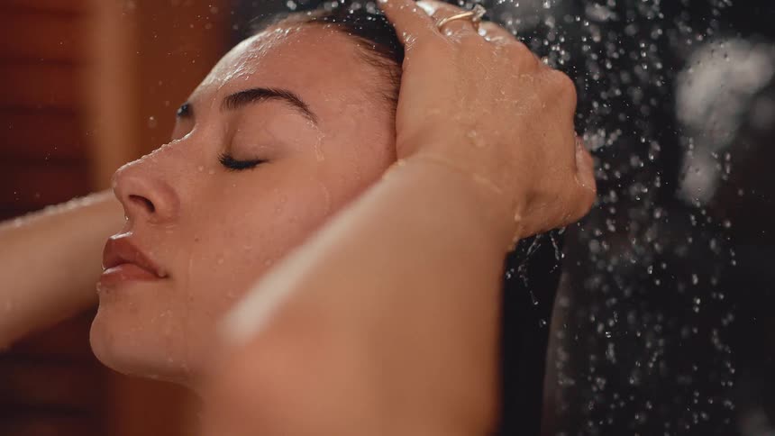 Serene woman with eyes closed washes her hair under a shower in a slow motion close-up