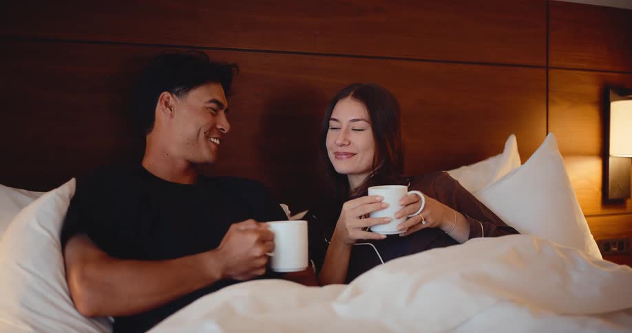 A happy young couple smiles and talks intimately while enjoying a warm beverage in bed at night