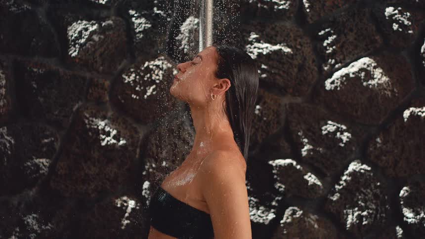 A serene woman in a black bikini top relaxes with her eyes closed under a refreshing shower against a dark stone wall