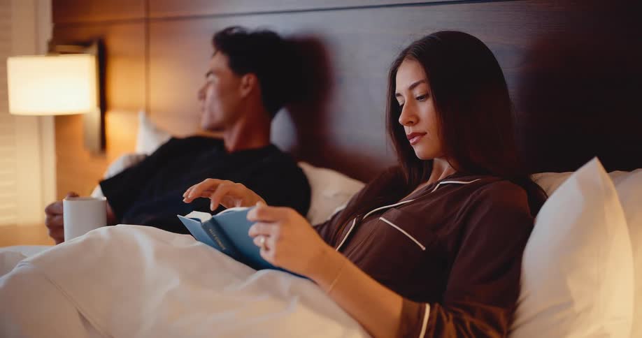A young woman enjoys a quiet evening reading a book in bed, while her partner relaxes with a drink beside her in a warmly lit room