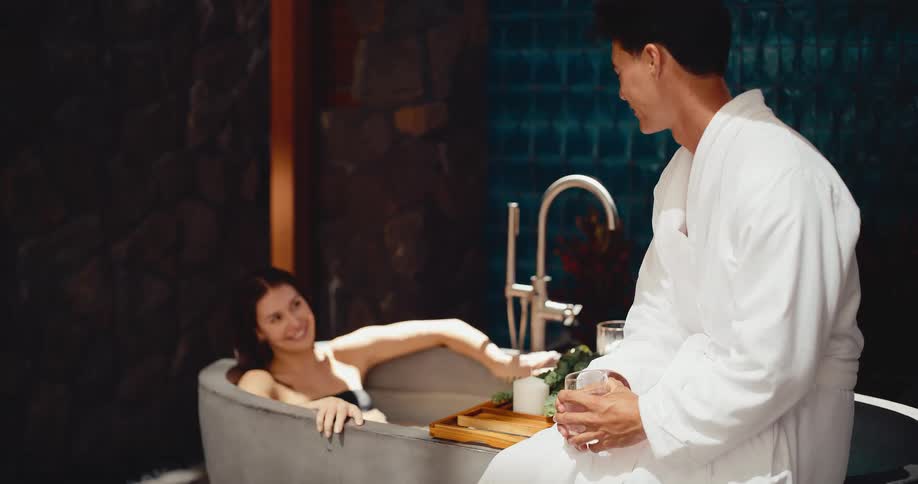 Slow motion shot of an attractive couple enjoying a romantic moment at the spa, with the woman in the bathtub and the man in a robe sitting beside her