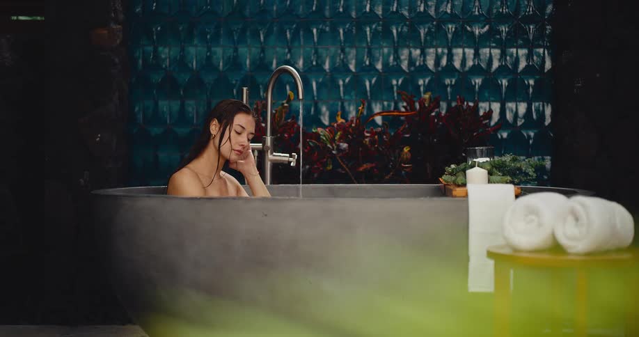 A beautiful young woman relaxes peacefully in a luxurious modern stone bathtub as it fills with water