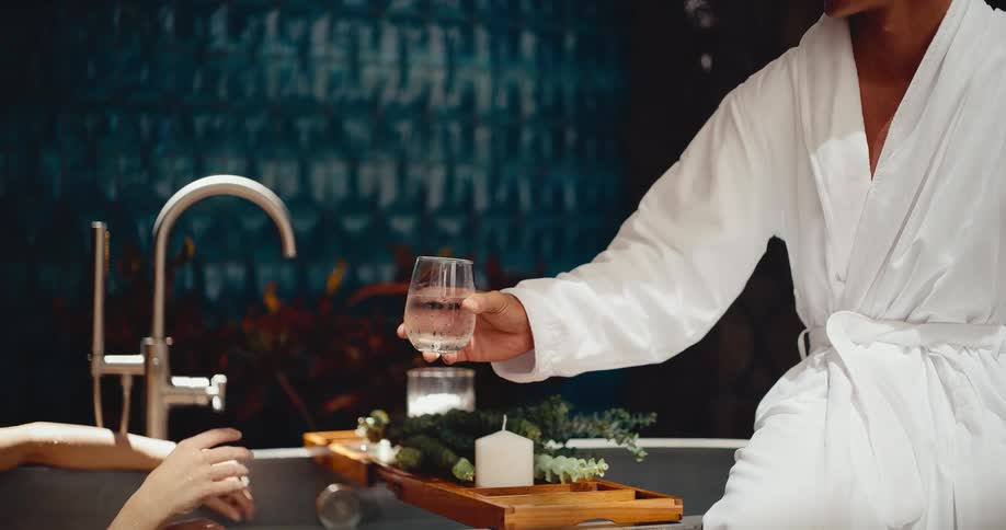 A man in a white bathrobe hands a glass of water to a smiling woman enjoying a relaxing bath in a luxurious spa setting