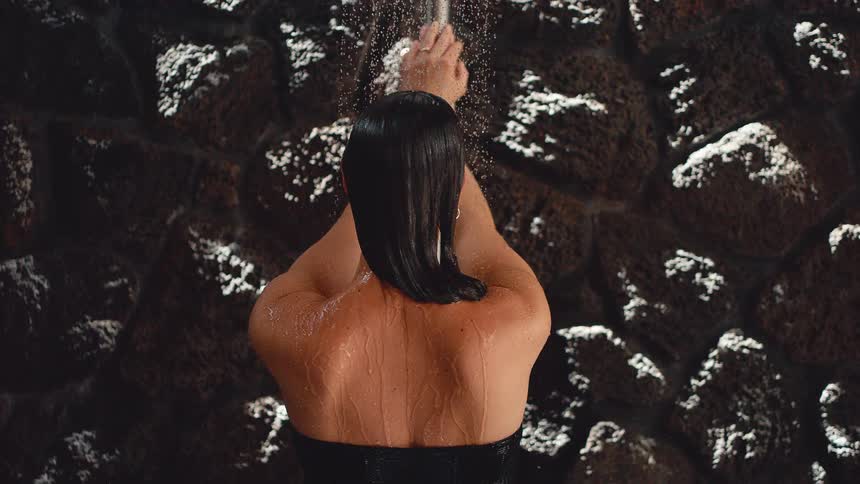 A woman with her back to the camera takes a relaxing shower against a dark stone wall in slow motion
