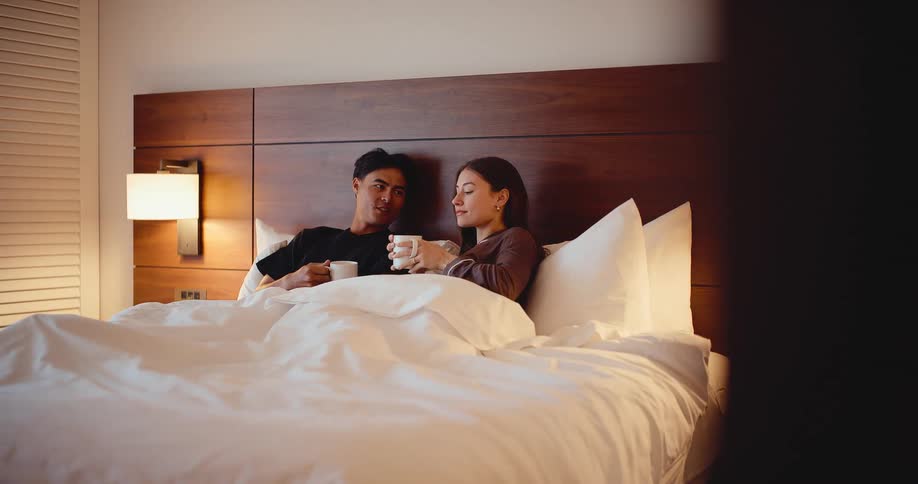Slow reveal shot of a happy young couple talking and drinking coffee together in bed in the morning