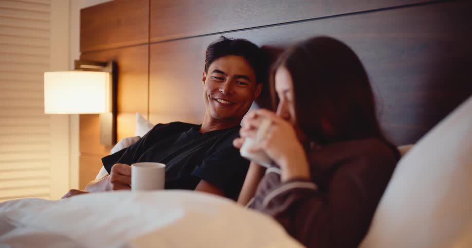 A young couple smiles and talks while enjoying a relaxing coffee in bed together in a hotel room