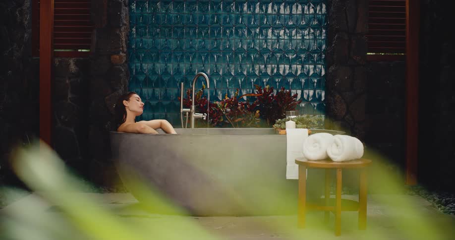 A woman peacefully relaxes in a modern stone bathtub in a luxurious outdoor spa setting with a tranquil waterfall feature