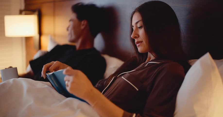 An attractive young woman reads a book in bed next to her partner who is relaxing with a warm drink