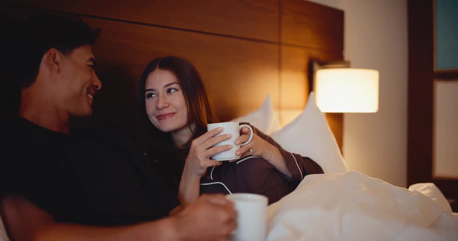 A happy young couple enjoys a quiet, romantic evening together, drinking from mugs and talking while relaxing in bed