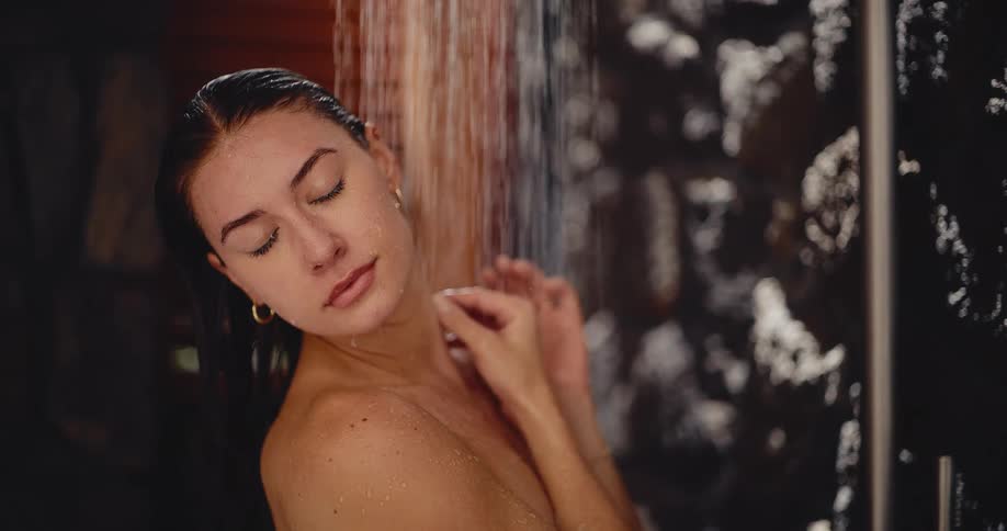 A beautiful young woman peacefully enjoys a warm shower, closing her eyes in a moment of tranquil self-care in slow motion