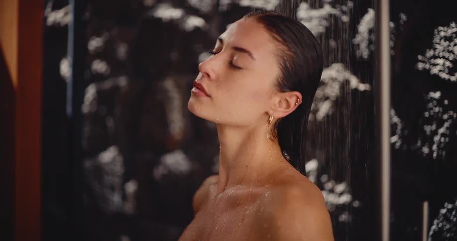 A serene woman with wet hair enjoys a relaxing moment under the warm water of a shower in slow motion