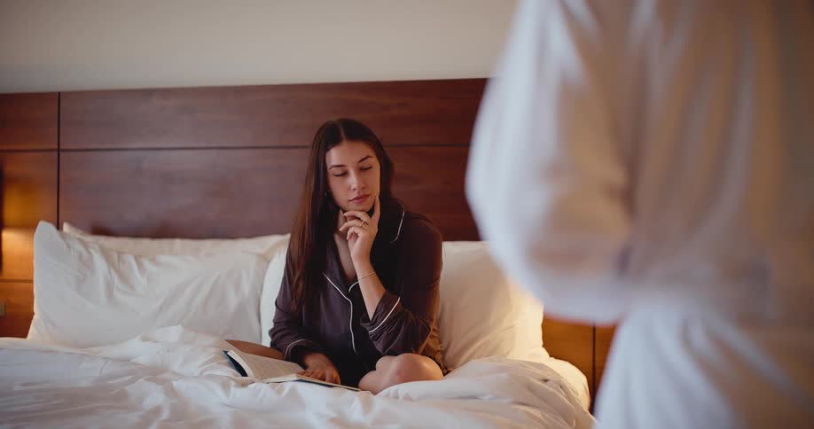 A young woman in pajamas reading a book in bed looks up and smiles lovingly as her partner approaches with a drink