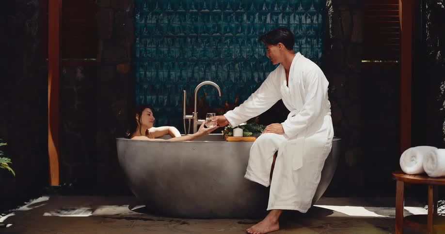 A man in a white robe hands a glass of wine to his partner as she relaxes in a luxurious modern bathtub