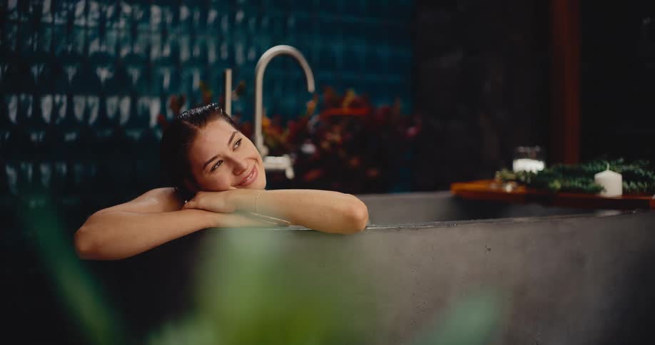 A young woman smiles contentedly while relaxing in a luxurious stone bathtub in a tranquil, plant-filled spa setting