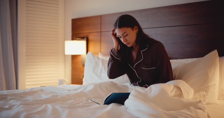 A young woman in her pajamas reads a book while sitting in a cozy bed at night