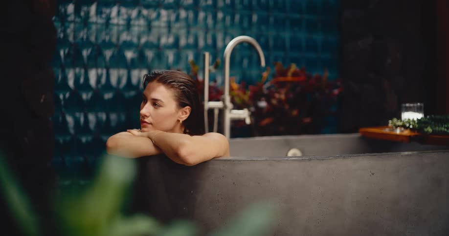 A serene young woman peacefully relaxes in a luxurious, modern stone bathtub, surrounded by lush plants