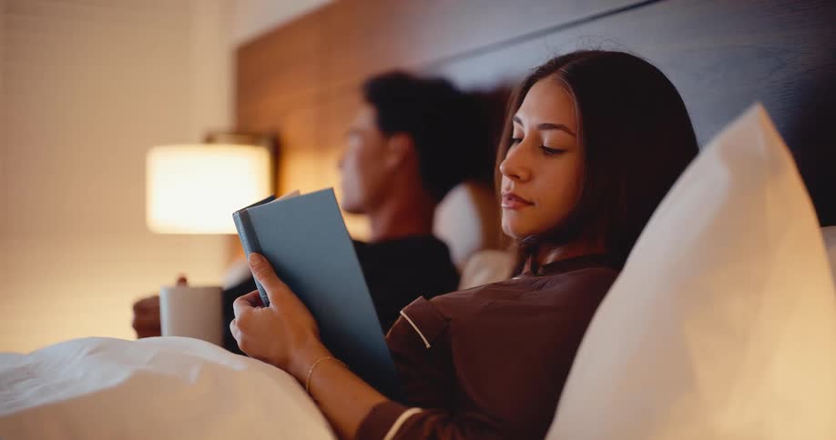 A beautiful young woman in pajamas reads a book in a cozy bed at night, with her partner relaxing in the background