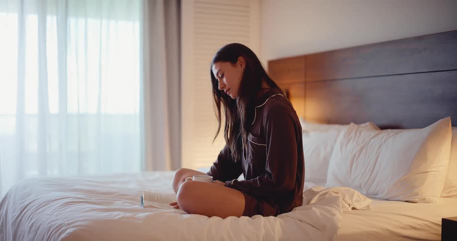 A relaxed young woman in brown pajamas sits on a white bed, reading a book and holding a mug in a softly lit bedroom