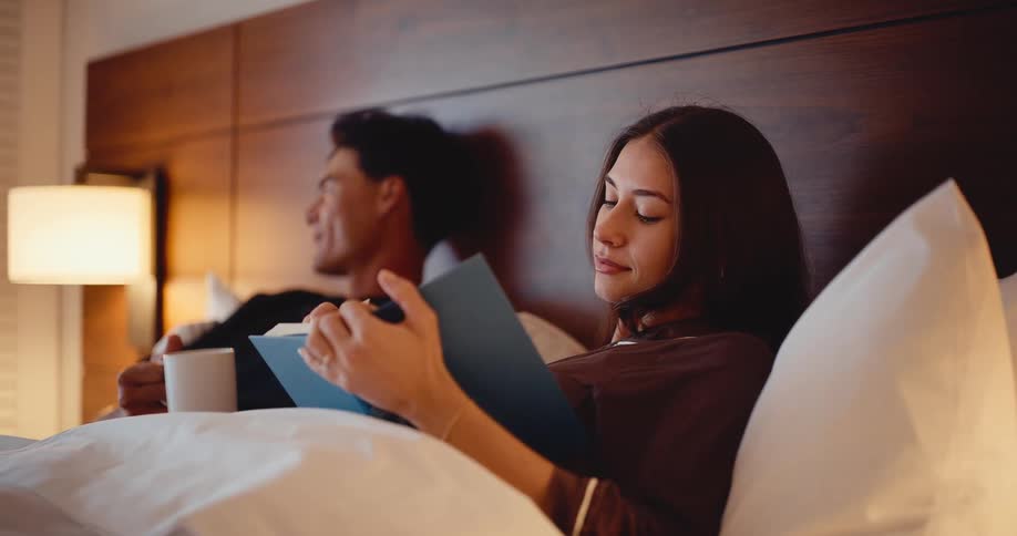 A relaxed couple enjoys a quiet evening in bed, with the woman reading a book and the man holding a mug
