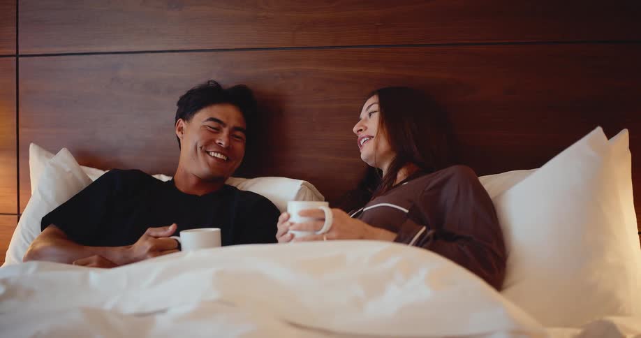Slow motion shot of a happy young couple laughing and enjoying their morning coffee together in bed