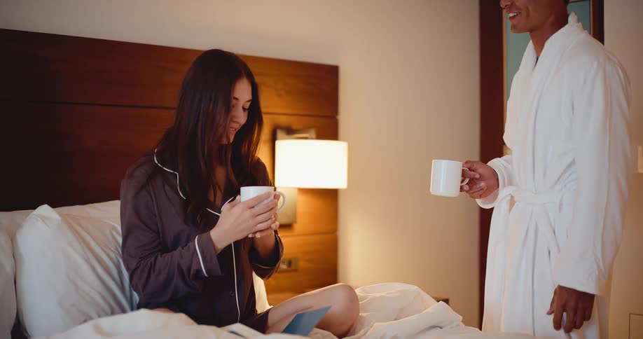 Slow motion shot of a man in a bathrobe bringing his smiling partner a warm cup of coffee in bed