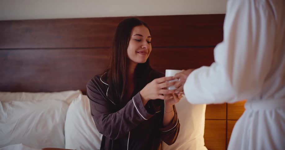 A young woman in pajamas reading a book in bed smiles happily as her partner brings her a warm cup of coffee in the morning