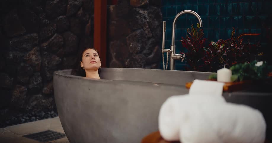 A beautiful young woman relaxes and closes her eyes while taking a luxurious bath in a modern stone tub at a spa