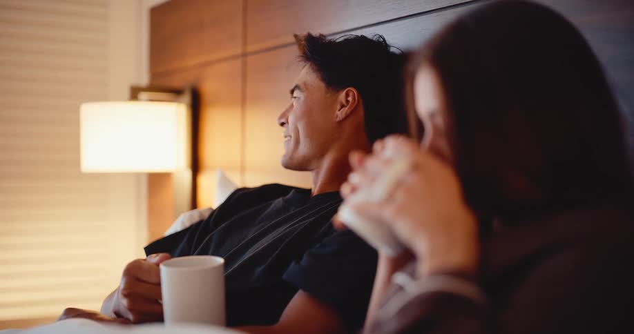 A smiling man looks lovingly at his partner as they relax and drink coffee together in a cozy bed