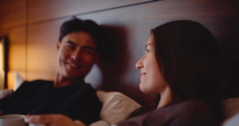 A romantic couple shares a quiet moment together, talking and smiling while having a drink in bed