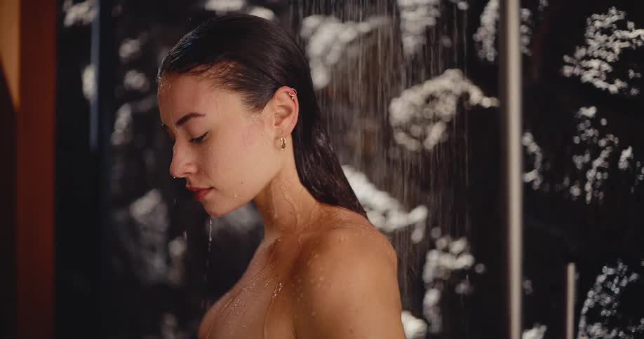 A serene young woman with eyes closed enjoys a relaxing and sensual shower in a modern, dark bathroom
