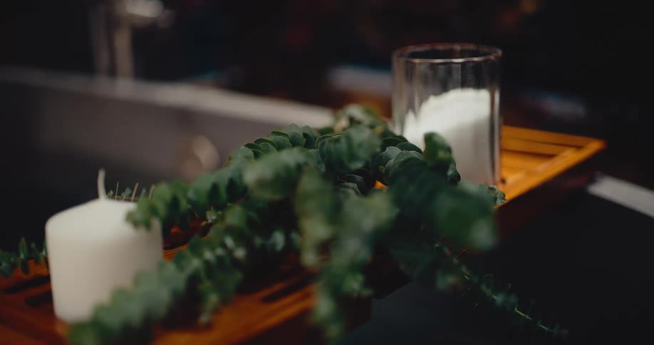 A close-up of a tranquil spa setting featuring a candle, eucalyptus, and bath salts resting on a wooden caddy over a bathtub