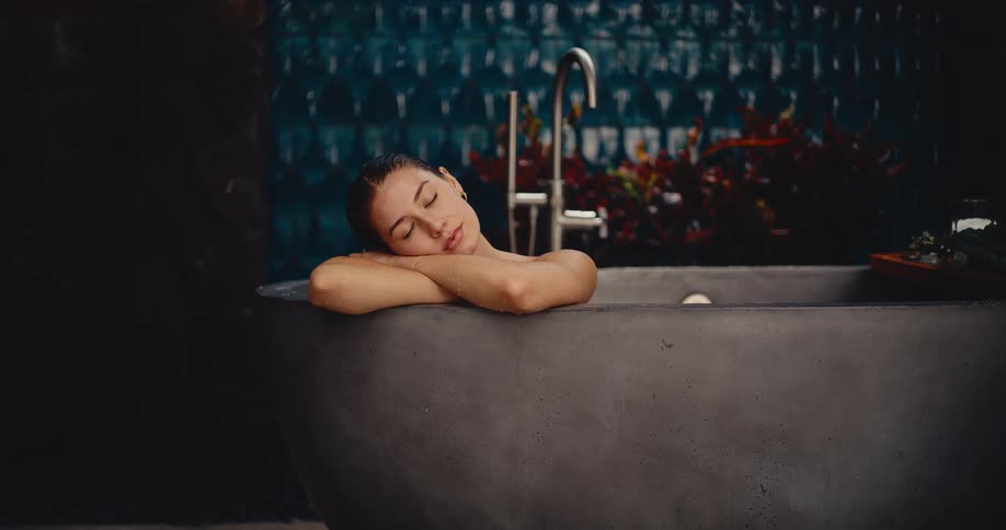 A beautiful young woman closes her eyes and relaxes peacefully while soaking in a luxurious, modern stone bathtub