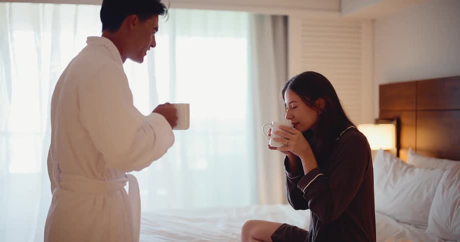 A loving husband brings his smiling wife a warm cup of coffee in bed in their sunlit hotel room