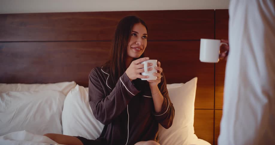 A thoughtful partner brings a warm morning drink to a beautiful woman reading a book in their cozy hotel bed