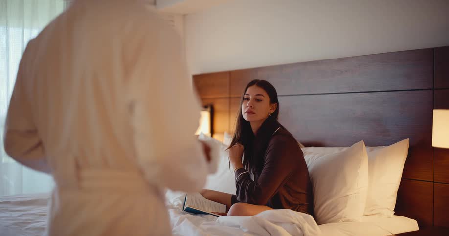 A woman in pajamas reads a book in a cozy bed while her partner in a bathrobe brings her a warm drink