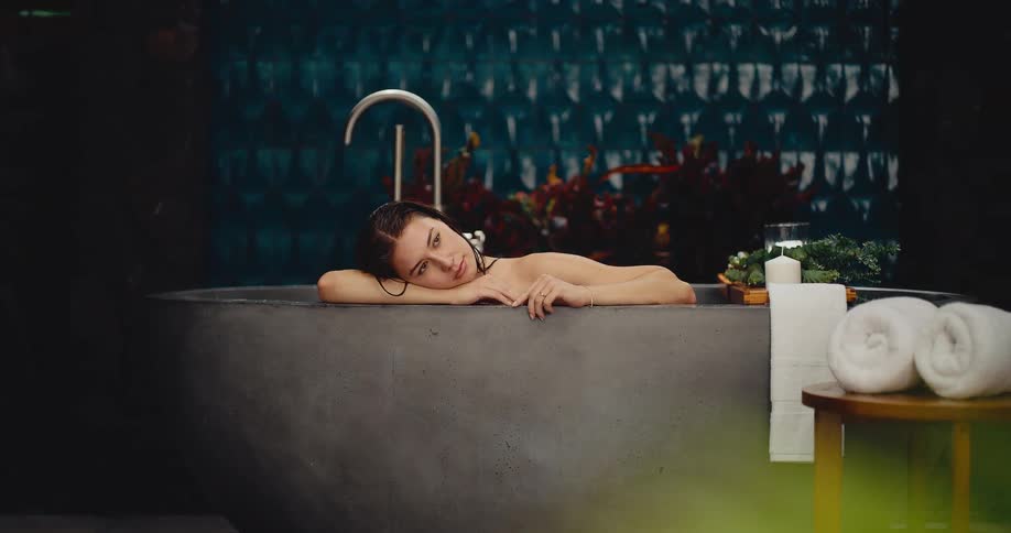 A beautiful woman with wet hair peacefully relaxes in a modern stone bathtub before opening her eyes