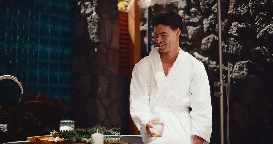 A handsome man in a white bathrobe smiles while enjoying a luxurious spa day by a running bath