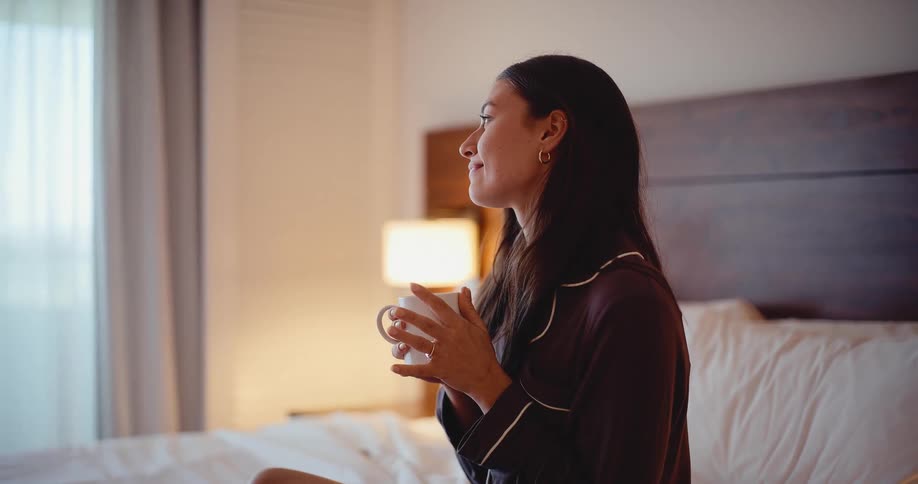 A young woman in pajamas enjoys her morning coffee while sitting on her bed in a hotel room