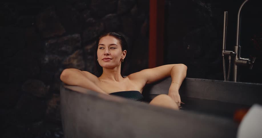 A beautiful young woman relaxes and smiles peacefully while soaking in a modern, stylish stone bathtub at a luxury spa