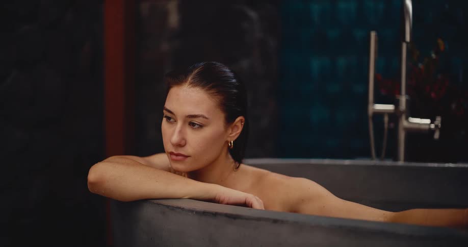 A thoughtful young woman with wet hair relaxes in a modern, stylish concrete bathtub, lost in thought
