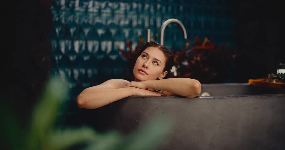 A beautiful young woman relaxes thoughtfully in a modern stone bathtub in a tranquil, plant-filled bathroom
