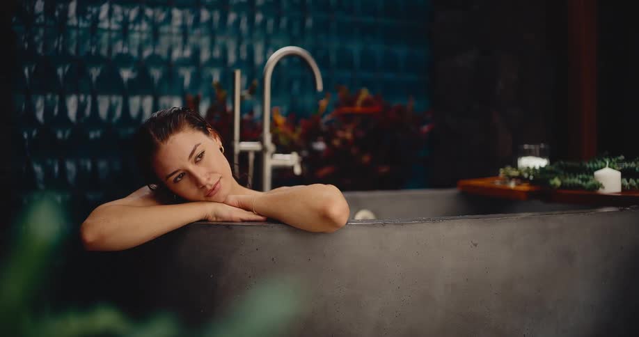 A pensive young woman relaxes in a modern stone bathtub before smiling peacefully and closing her eyes in a luxurious spa setting