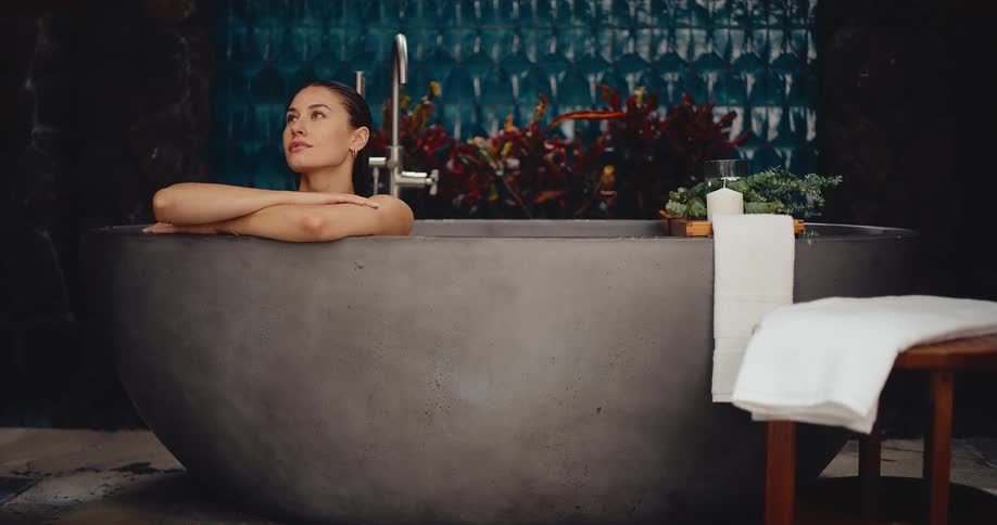 An attractive woman takes a moment for self-care, relaxing in a large stone bathtub in a modern, spa-like bathroom