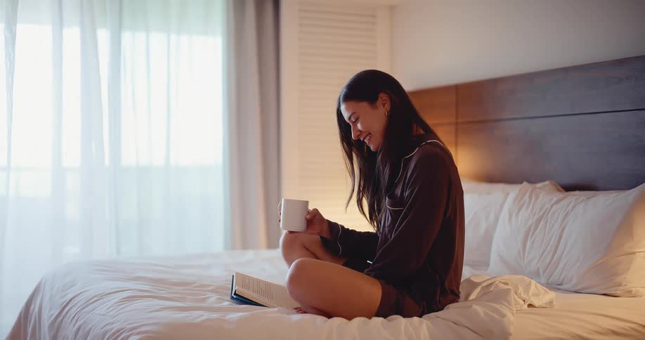 Happy young woman in pajamas enjoys a relaxing moment with a book and a warm drink while sitting on a cozy bed