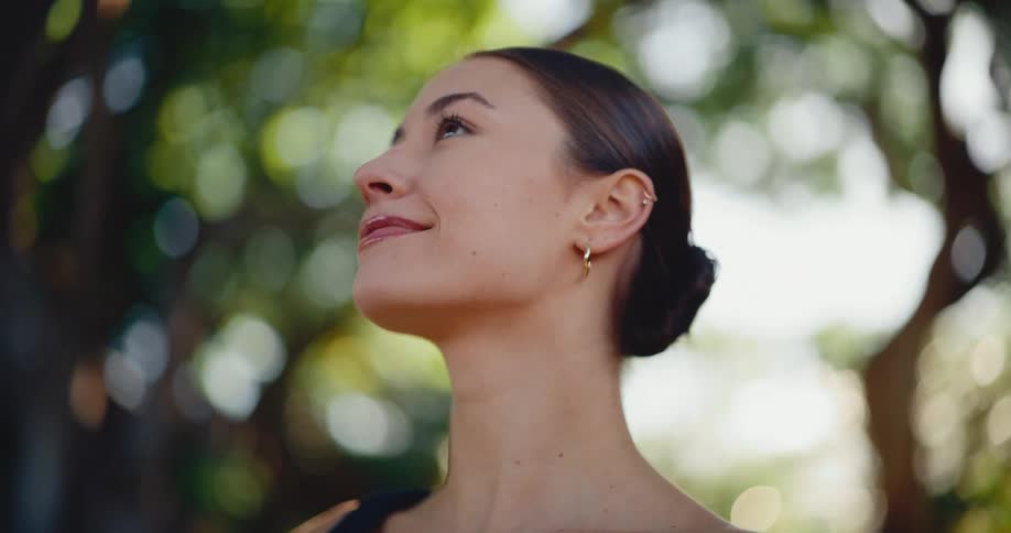 A serene young woman with her hair in a bun looks up peacefully while standing in a lush green park